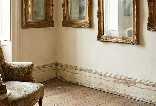 Состаренный белый деревянный плинтус с потёртой краской вдоль стены, старинное кресло с цветочной обивкой