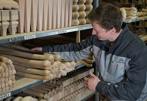 Мастер выбирает деревянные заготовки на стеллаже, демонстрируя разнообразие форм и пород древесины для изготовления стульев.