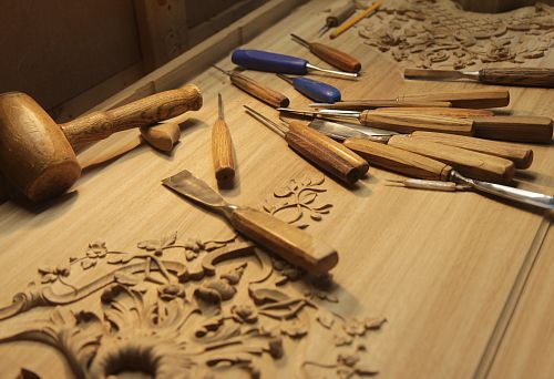 Workbench with a set of chisels and a wooden mallet, next to partially carved patterns on solid wood, illustrating the wood processing process.
