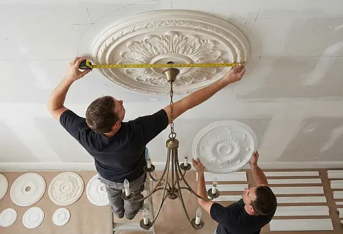 Polyurethane ceiling molding for a chandelier &mdash; a master measures the diameter of a ceiling rosette with a tape measure, a second holds a sample of a smaller rosette