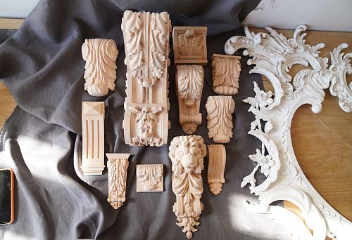 Carved wooden appliqu&eacute;s and decorative furniture elements, including lion heads, leaves, and floral ornamentation, mounted on fabric and prepared for decorative painting.