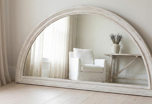 An arched mirror in a wide white frame with a distressed wood effect stands on the floor against the wall; the reflection shows light curtains, a white armchair, and a console table in a calm Scandinavian interior
