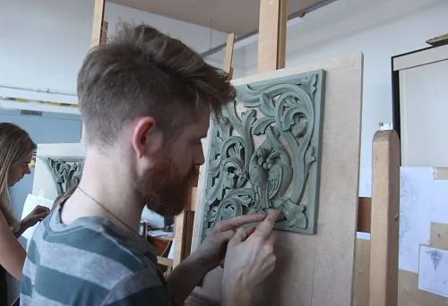 The artisan hand-carves a relief with an owl and plant ornament on a board according to an original sketch in the workshop.
