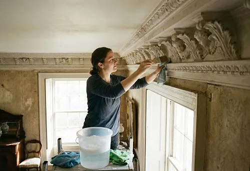 A woman carefully cleans a relief ceiling cornice using a brush and cloth; a container with water stands nearby, the interior looks antique, with rich decorative molding