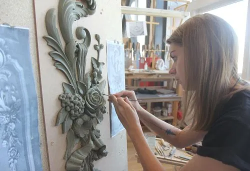 Artist hand-carves a relief with flowers and grapes according to a sketch in the workshop, using a fine tool to refine details.
