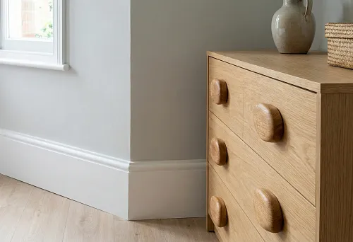 Light-colored MDF baseboard and solid wood furniture handles on a chest of drawers, an example of contrast between budget finishing and accent elements in an interior.