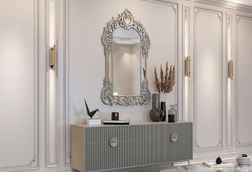 Interior with a modern natural wood chest and a large mirror in an ornate classical frame on a light wall with decorative moldings.