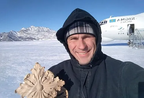 A man in winter clothing holds a carved wooden decorative element against a snowy landscape