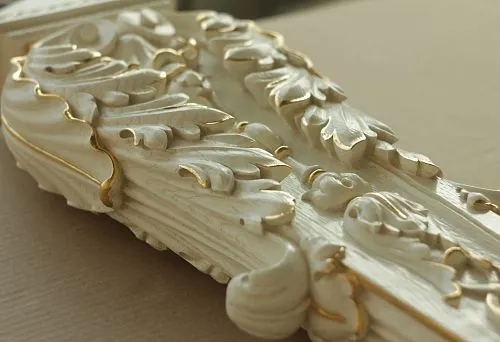 Decorative carved element for staircase post with white finish and gold patina