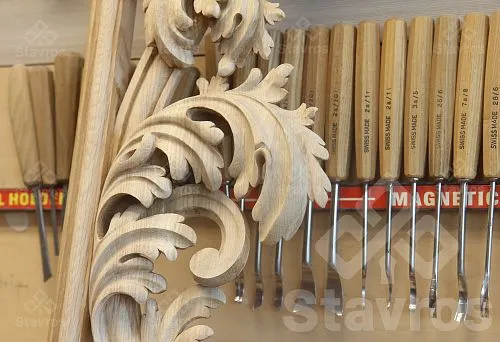 A large carved wooden element with acanthus leaves against a backdrop of professional chisels in a carpentry workshop.