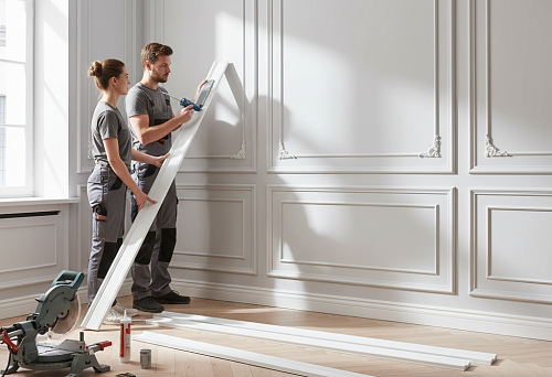 Craftsmen install polyurethane moldings on a wall in a light interior, demonstrating the installation process and neat finishing of decorative panels.
