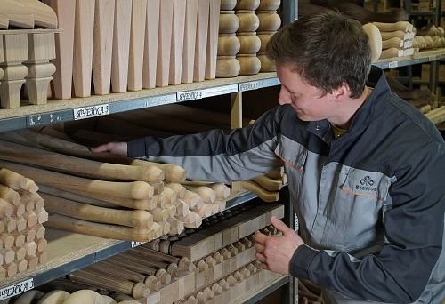 The craftsman selects wooden blanks on a shelf, demonstrating the variety of shapes and wood species for making chairs.