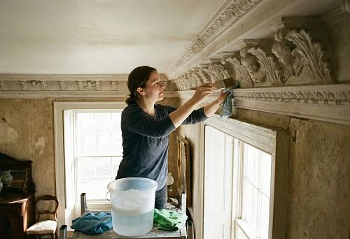 A woman carefully cleans a relief ceiling cornice using a brush and cloth; a container with water stands nearby, the interior looks antique, with rich decorative molding