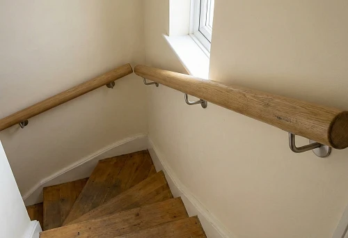 Wall-mounted round wooden handrail on a narrow staircase with a turn, mounted on metal brackets, light walls.