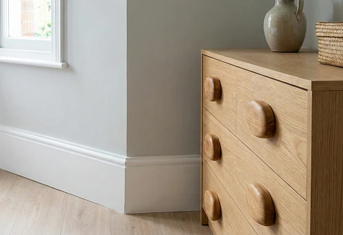 Light-colored MDF baseboard and solid wood furniture handles on a chest of drawers, an example of contrast between budget finishing and accent elements in an interior.