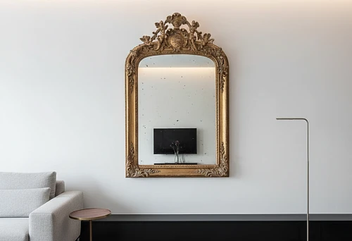 Antique mirror in a carved gold frame on a white wall above a wide black baseboard, minimalist living room with a sofa and floor lamp