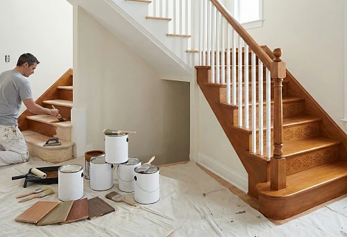 Painting a wooden staircase: a master applies interior paint to steps with a brush, nearby paint cans, tools, and shade samples
