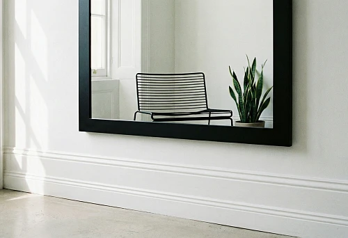 Black rectangular mirror in a thin frame on a light wall above a tall white baseboard; sunbeams on the wall