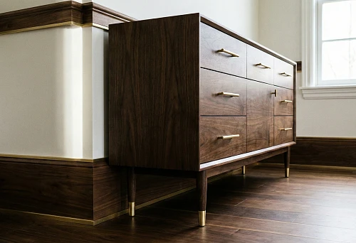 Dark wood chest of drawers with metal-colored furniture handles and supports: the baseboard and molding around the perimeter of the walls form a single finishing line.