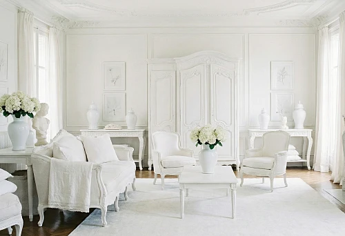 Bright living room with white classic furniture, fireplace, wall panels, molding, white carpet, and vases with hydrangeas