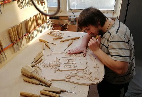 The master manually carves heraldic relief from wood using chisels in the STAVROS workshop.