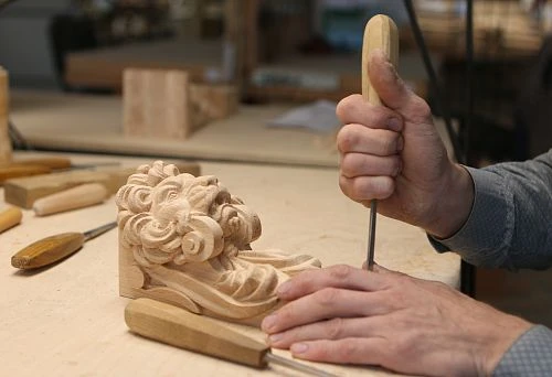 The master's hands using a chisel for wood carving create a detail with an artistic lion's head.